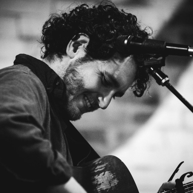 Sasha singing into a microphone with an acoustic guitar, black and white