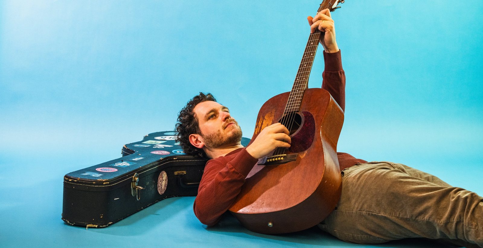 Sasha lying on his back holding an acoustic guitar against a sky-blue studio backdrop