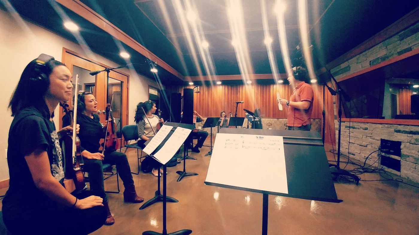 A string section at music stands in a wood-paneled recording studio, Sasha at the far end